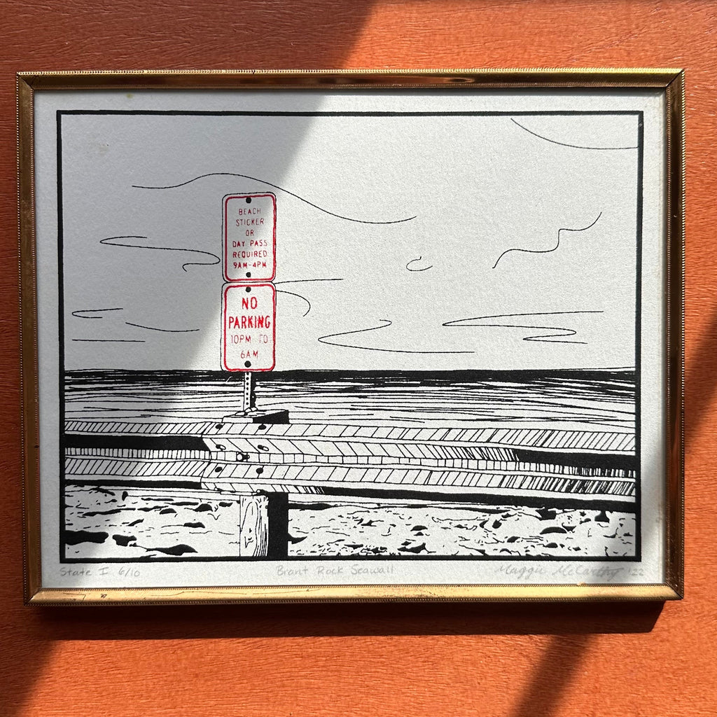 Framed black and white artwork of a beach scene with a sign on a wooden surface
