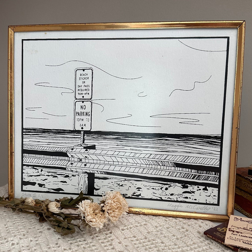 Framed black and white drawing of an ocean landscape with a gold frame on a textured surface.