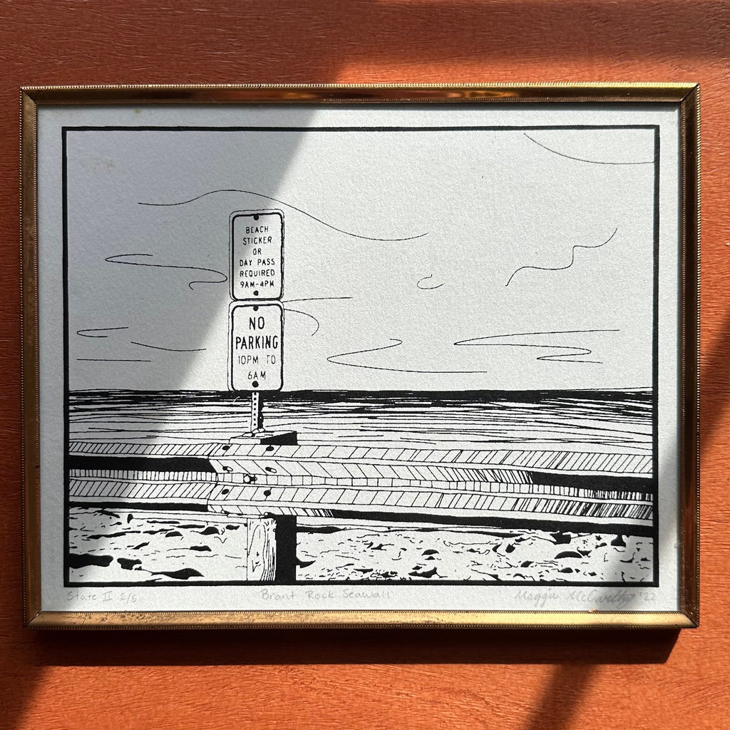 Framed black and white drawing of an ocean landscape with a sign on an wooden table.