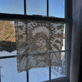 Decorative lace curtain hanging in front of a window with blue sky and trees outside.