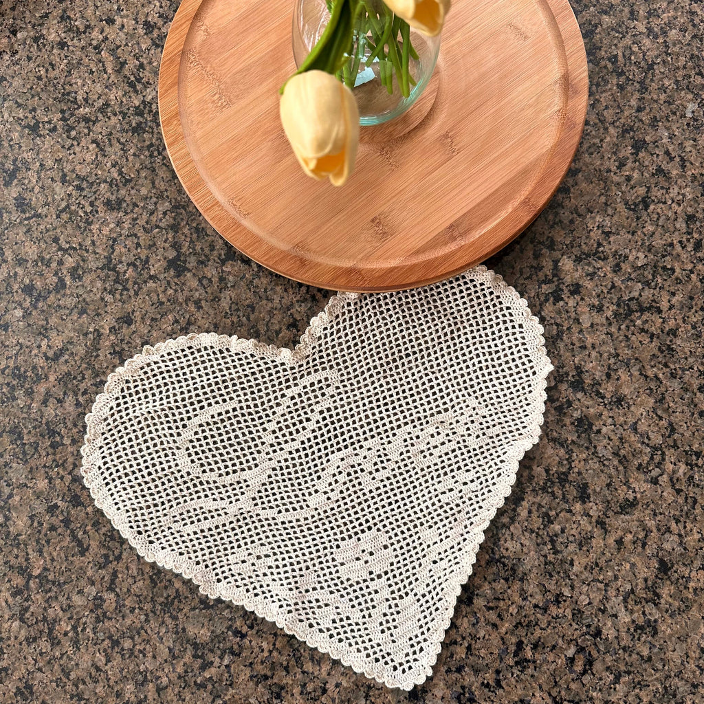 a heart-shaped crochet doily is seen on a granite table with a wooden plate and flowers in a vase above