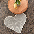 a heart-shaped crochet doily is seen on a granite table with a wooden plate and flowers in a vase above