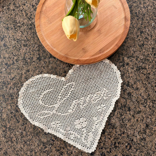 a heart-shaped crochet doily is seen on a granite table with a wooden plate and flowers in a vase above