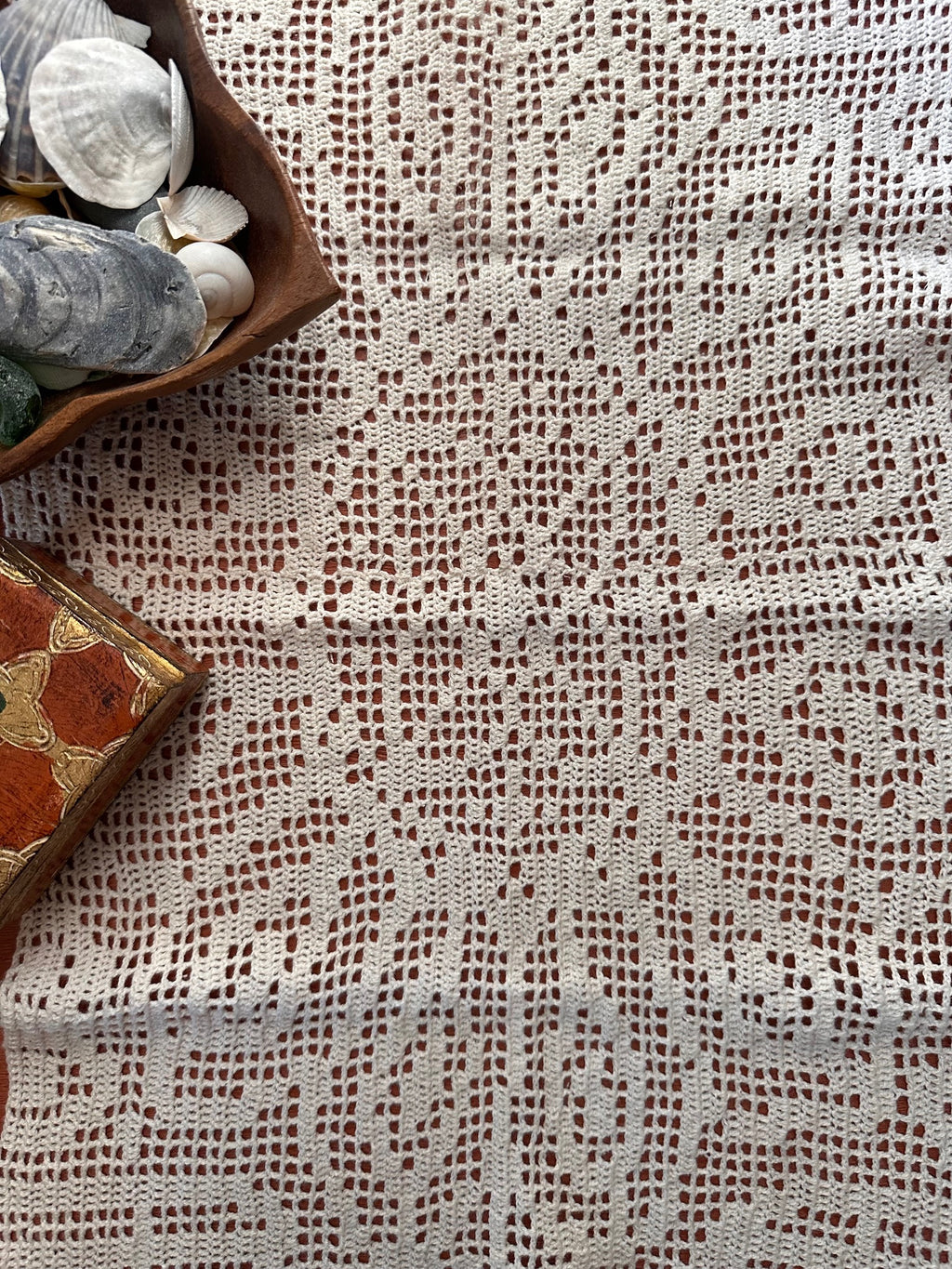 Close-up of a filet crochet textile with a bowl of shells and decorative wooden box on top